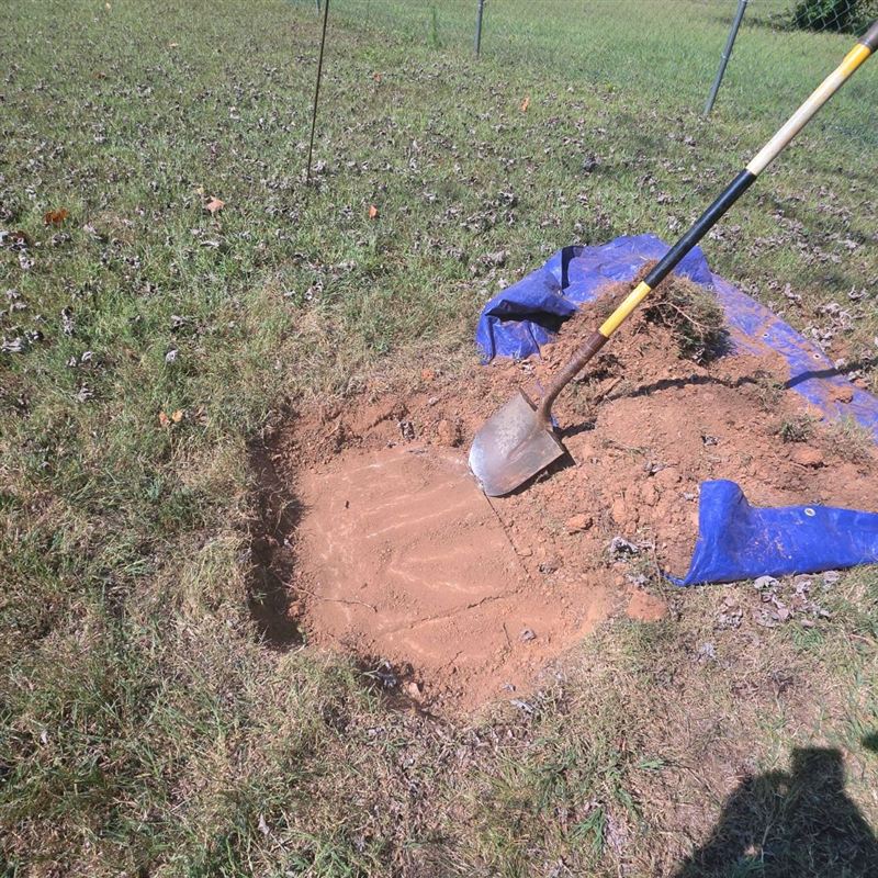 Shovel in freshly dug red clay on a blue tarp beside a shallow hole over a buried septic tank lid